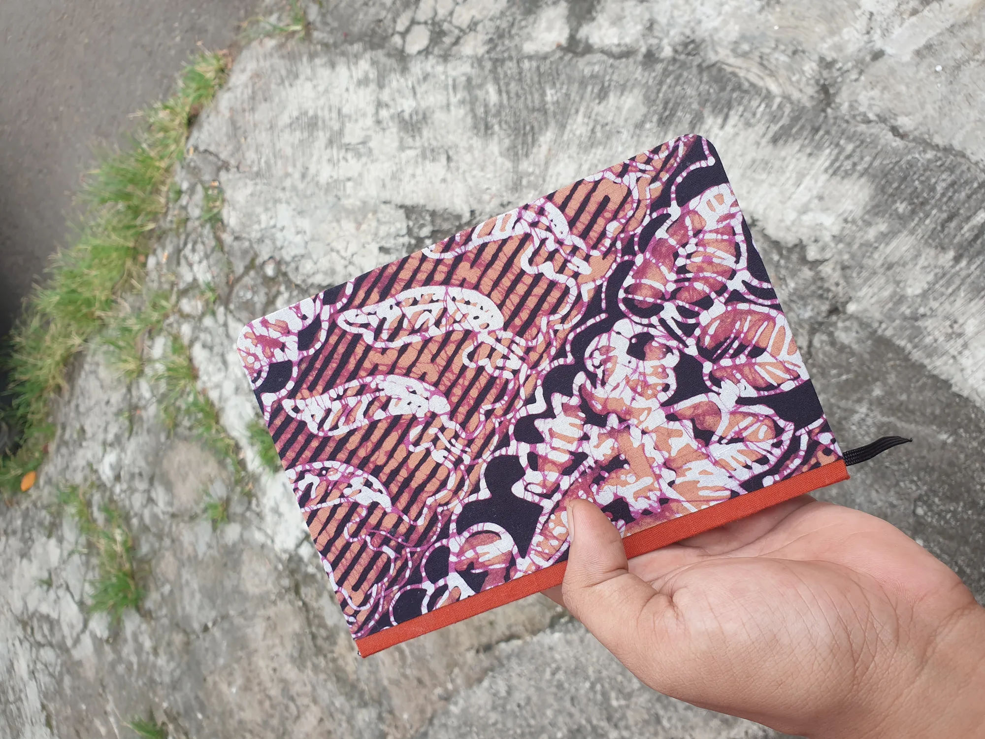 Buku catatan bersampul kain batik merah dan hitam, dipegang tangan di atas batu berlumut