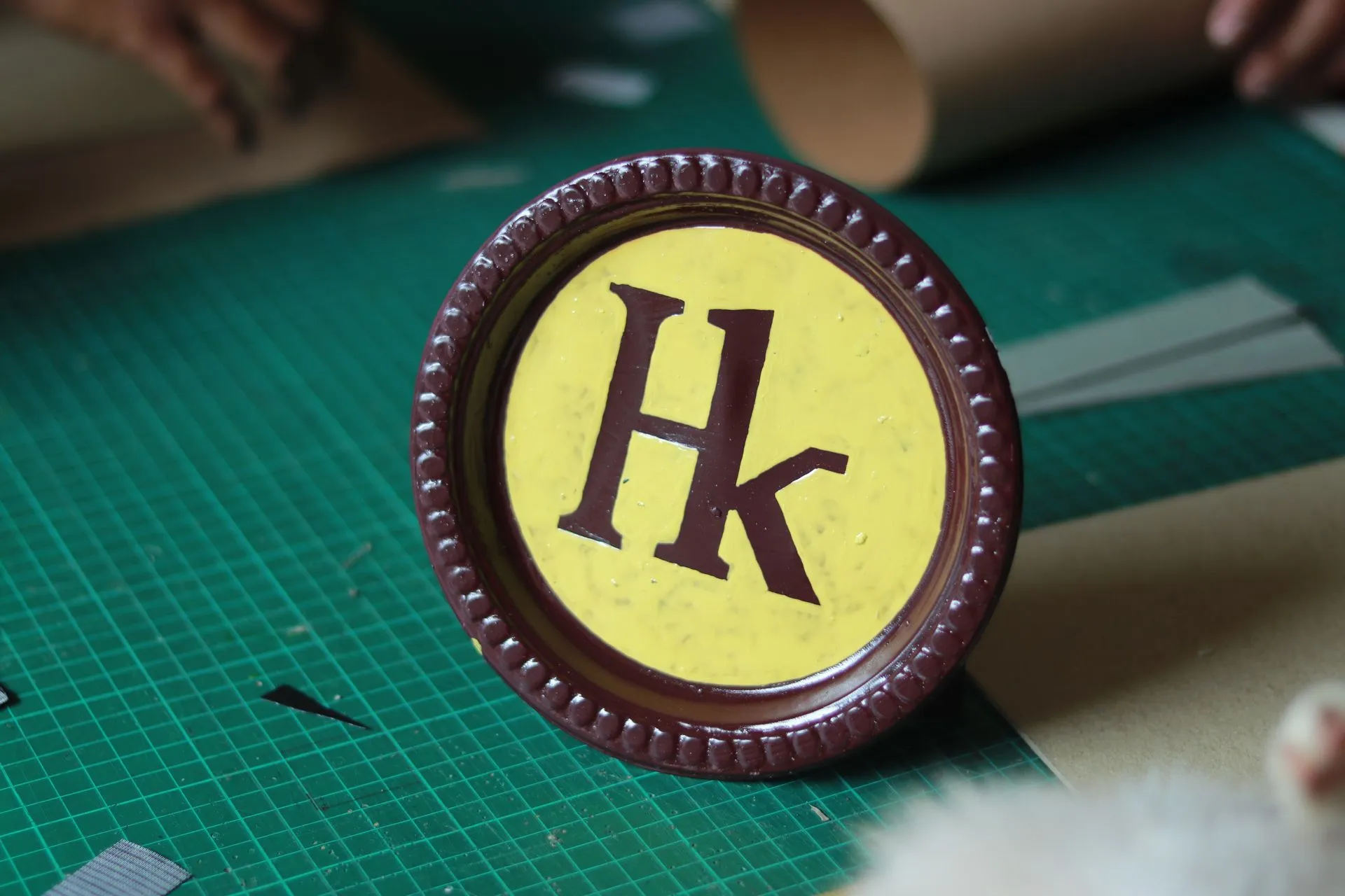 Monogram Hk dicat tangan pada medali kayu bundar, di atas alas potong hijau, dikerjakan oleh perajin mitra
