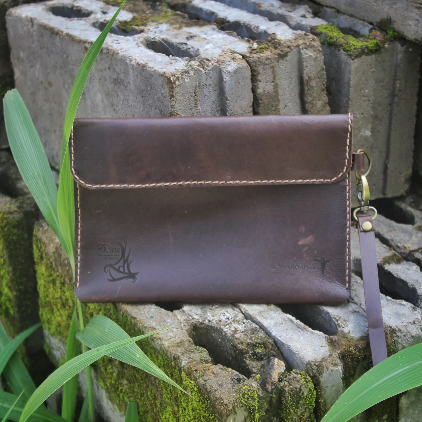 A brown leather clutch with a wrist strap placed on a stack of mossy concrete bricks.
