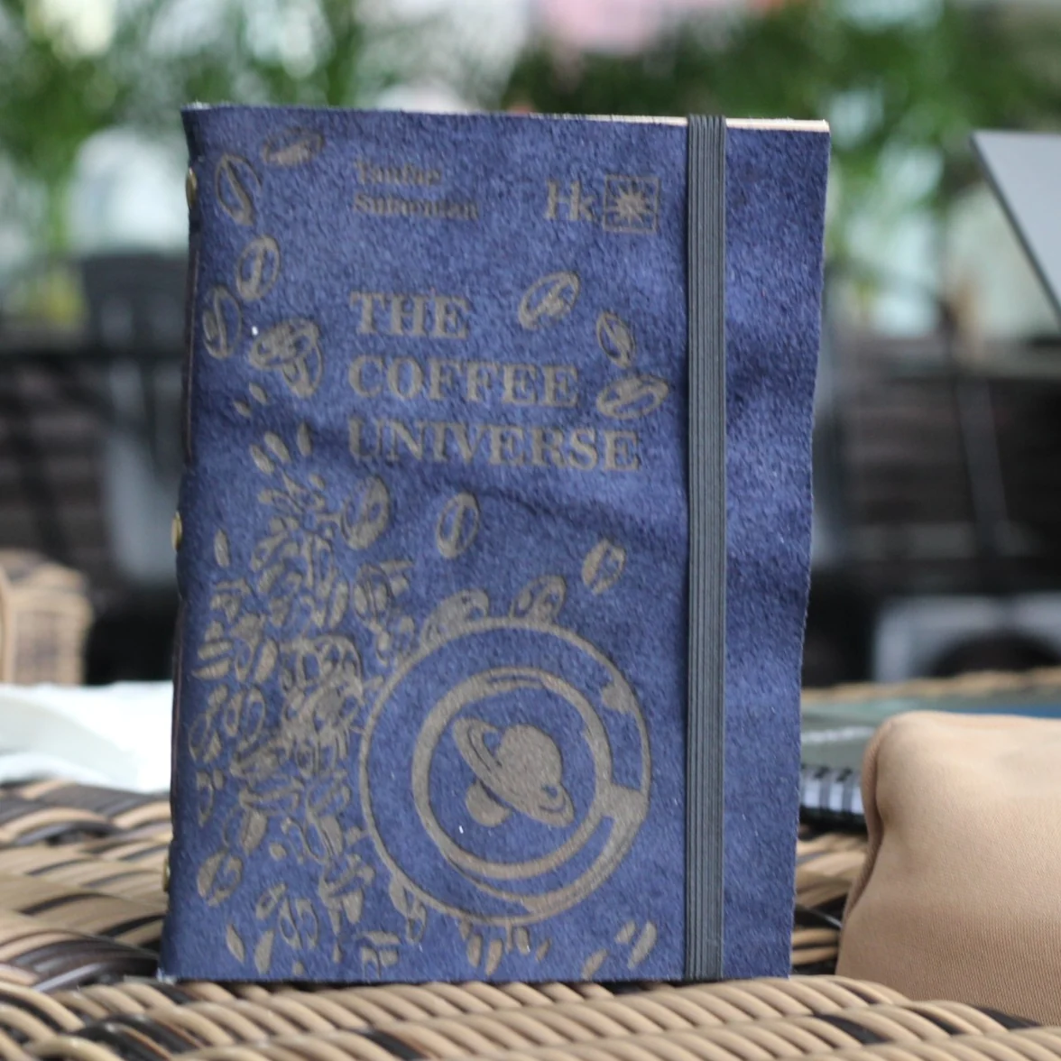 A blue suede leather journal with the coffee universe text on a woven table.