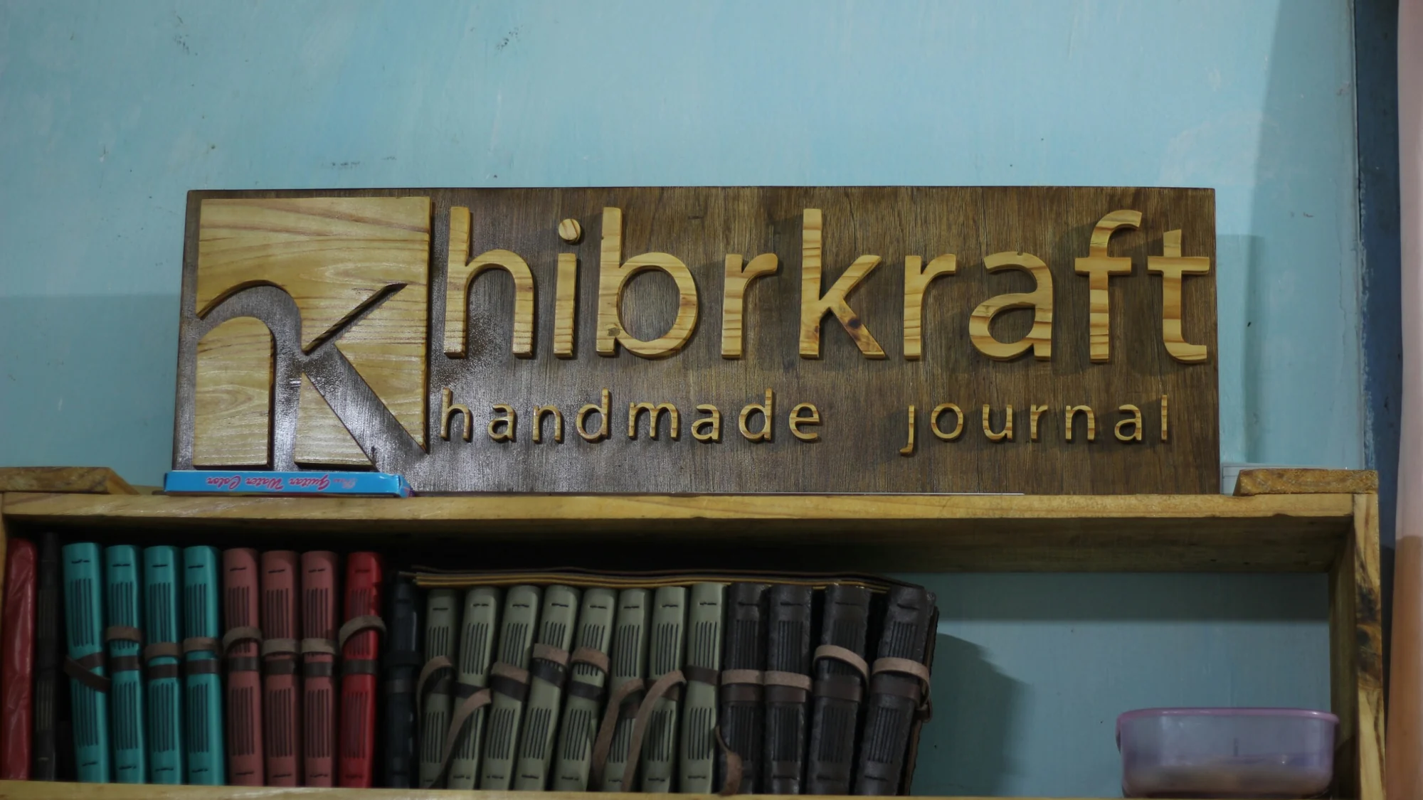 Papan kayu bertuliskan Hibrkraft di atas rak berisi jurnal kulit