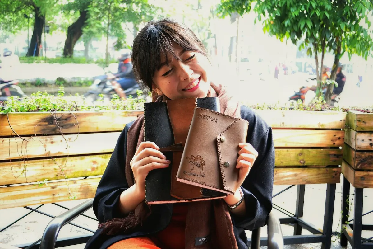 Anya Hasna duduk di bangku taman memegang dua jurnal kulit Hibrkraft, yang bersampul tan bercap gambar buatannya sendiri