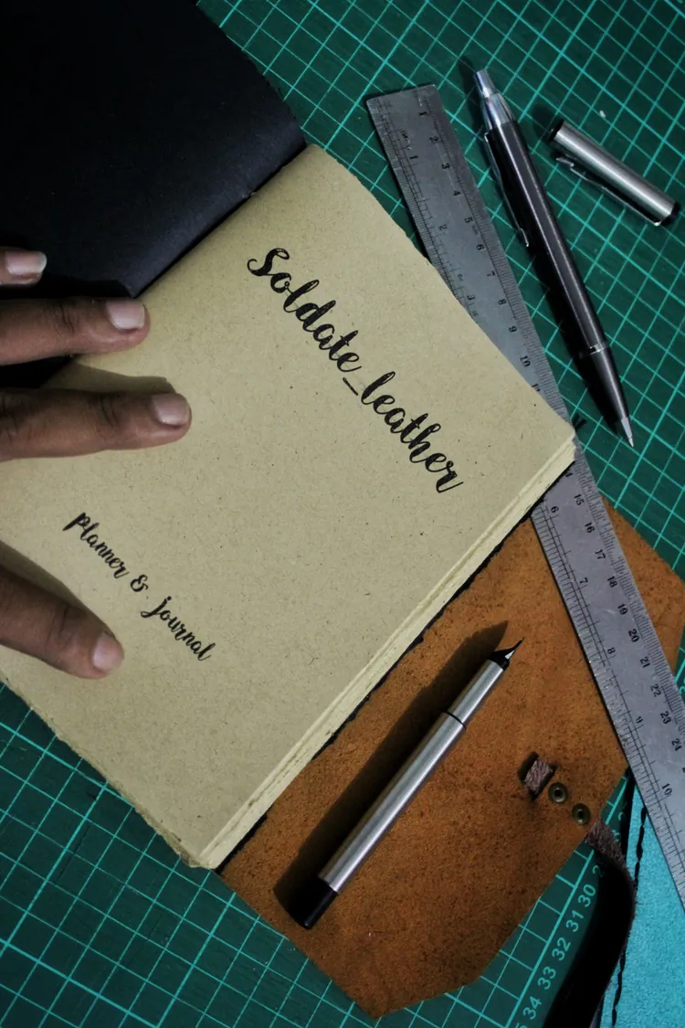 Sampul jurnal kertas kraft bertuliskan tangan Soldate_leather, Planner & Journal, di atas matras potong hijau bersama penggaris baja dan dua pena teknis
