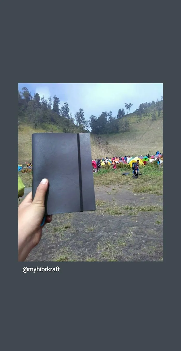 Story Instagram: jurnal bersampul gelap dipegang di depan lapangan kemah penuh tenda warna-warni