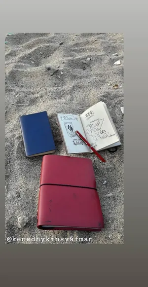 Story Instagram: jurnal biru, buku sketsa terbuka dan jurnal kulit merah tergeletak di atas pasir