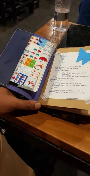 Tangan memegang jurnal terbuka yang penuh stiker dan catatan tulisan tangan di meja kafe