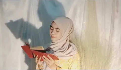 Perempuan berhijab membaca jurnal bersampul merah di depan tirai putih