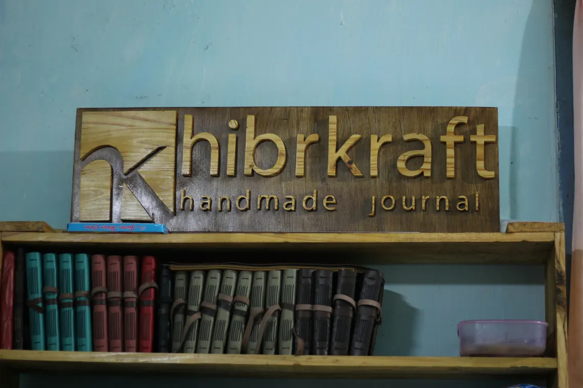 Papan kayu bertuliskan nama Hibrkraft di atas rak jurnal kulit