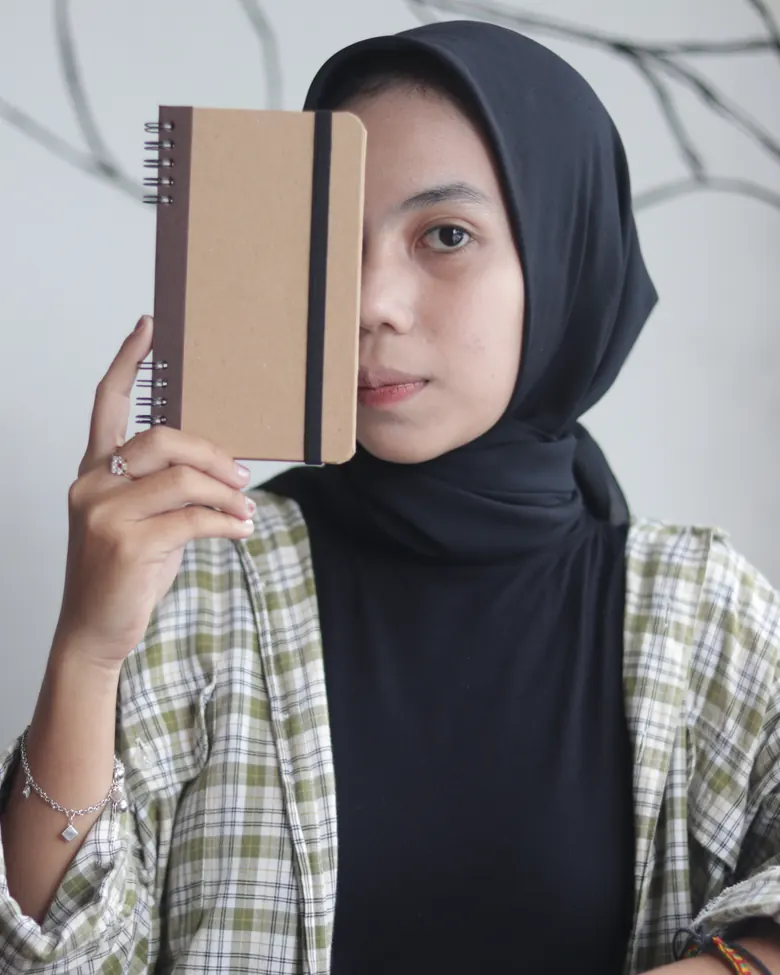 Seorang perempuan memegang buku catatan spiral di samping wajahnya